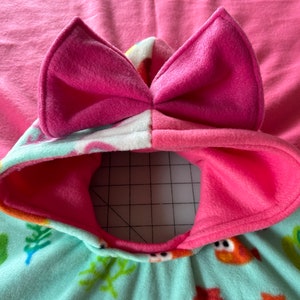 May include: A pink hooded fleece blanket with a bow on the hood. The blanket is lined with a white and blue fabric with a seahorse and seaweed pattern.