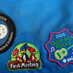 May include: A blue fabric patch with three embroidered patches. The top patch is black with a yellow crescent moon and a white moon with craters. The text "e Day 2024" and "outs of Central Indiana" are on the patch. The middle patch is green with the text "First Meeting" and three cartoon birds. The bottom patch is blue with the text "2024 CAMP ADA ROCKS" and a green guitar with music notes. The text "gsci" is on the patch.