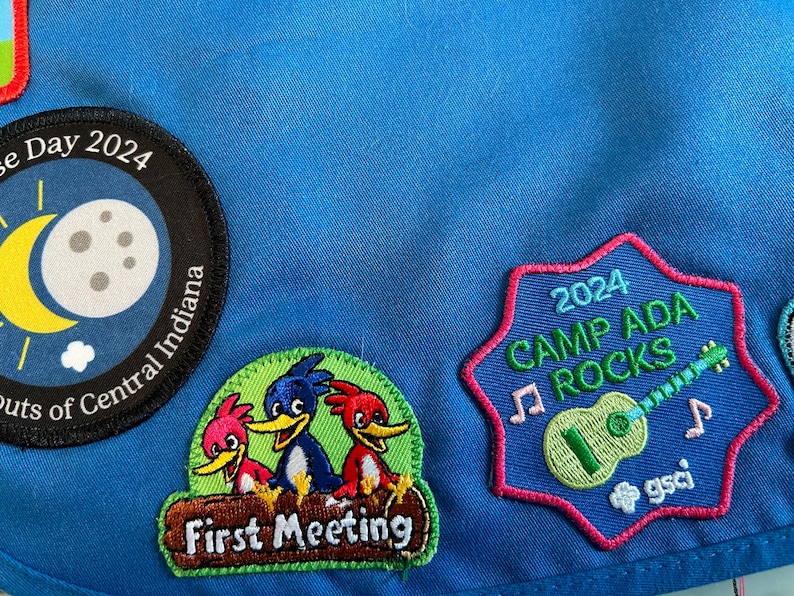 May include: A blue fabric patch with three embroidered patches. The top patch is black with a yellow crescent moon and a white moon with craters. The text "e Day 2024" and "outs of Central Indiana" are on the patch. The middle patch is green with the text "First Meeting" and three cartoon birds. The bottom patch is blue with the text "2024 CAMP ADA ROCKS" and a green guitar with music notes. The text "gsci" is on the patch.