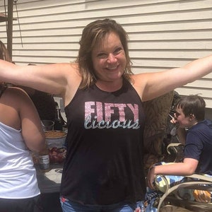 May include: A woman wearing a black tank top with pink glitter lettering that says "Fifty Licious".