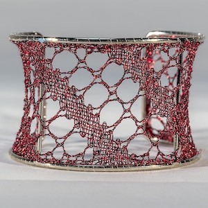 Pode incluir: Uma pulseira de punho prateada com um design de tecido aberto vermelho e preto. A pulseira tem uma faixa larga com um design delicado e rendado. O trabalho aberto permite vislumbrar o fundo. Um acessório elegante.