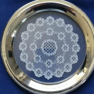 May include: A silver serving tray with a beaded edge, displaying a white lace doily. The doily is centrally placed, offering a vintage feel. The background is a solid blue.
