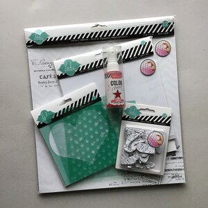 Könnte beinhalten: Ein Set Bastelbedarf, bestehend aus einer rosa Sprühflasche mit der Aufschrift "Color Shine", einer weißen Schablone in Herzform, einem Blatt weißem Karton und einer Packung weißer Papierverzierungen.
