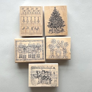 May include: Five wooden stamps with festive Christmas designs. The stamps depict reindeer, Christmas trees, snowmen, nutcrackers, a gingerbread house, and carol singers with the text "Season's Greetings". Ideal for seasonal crafts and card making.