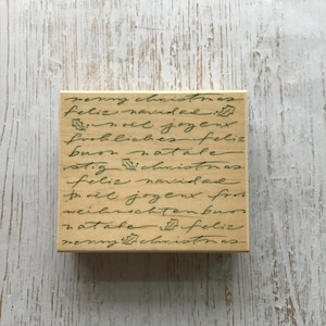 Op de afbeelding: Een houten stempel met de tekst "merry christmas, feliz navidad, noil joyeux, frohliches feliz, buon natale, stig christmas, feliz navidad, troil joyeux from, weihnachtenbuos, natale feliz, merry christmas" geschreven in cursief schrift. De tekst is in groene inkt en er zijn kleine groene hulstbladeren door de hele tekst.