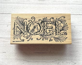 以下が含まれることがあります： 「NOEL」の文字が黒い装飾的なフォントで書かれた木製のスタンプ。文字は黒い植物のイラストと鳥のイラストで囲まれています。長方形のスタンプは明るい木材の背景に置かれています。