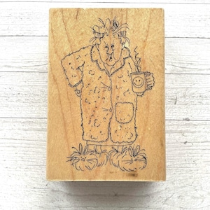 May include: A rectangular wooden rubber stamp featuring a black ink line drawing of a person with messy hair in curlers, wearing a textured robe and fuzzy slippers, holding a steaming mug with a smiley face design on the front of the mug.