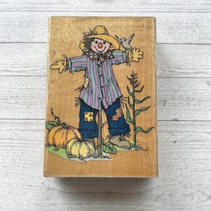 May include: A rectangular wooden rubber stamp featuring a colorful illustration of a scarecrow wearing a yellow hat, a purple striped shirt, and blue pants, standing next to two orange pumpkins and a green corn stalk on a light wood block.