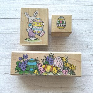 May include: Three wooden Easter stamps. One stamp features a bunny holding a decorated egg. Another shows a decorated egg. The third stamp has a design of Easter eggs, flowers, and grass.