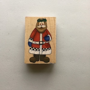 May include: A wooden stamp with a cartoon illustration of Santa Claus wearing a red and white suit, a green wreath, and holding a red heart.