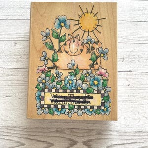 May include: Wooden stamp featuring a cartoon cat with a sun and flowers. The cat is orange and white, surrounded by blue and pink flowers. The stamp includes the text "Warm Thoughts, Warm Wishes..." in a decorative banner.