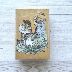 May include: A wooden stamp featuring a charming illustration of three kittens playing in a garden. One kitten is reaching for a butterfly, while the others are interacting with a blanket. The artwork is in soft pastel colors.