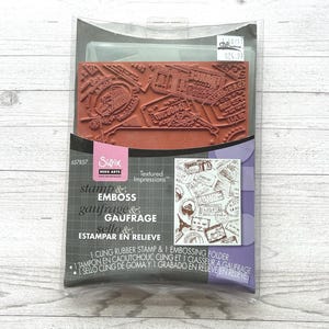 May include: A Sizzix Hero Arts stamp and emboss set in clear plastic packaging. The set includes a rectangular terracotta-coloured rubber stamp featuring travel-themed postage stamps and a clear plastic embossing folder with a matching design.
