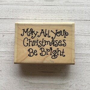 Op de afbeelding: Een houten stempel met de tekst "May All Your Christmases Be Bright" in een speels lettertype.