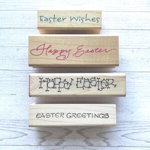May include: Four wooden Easter stamps with text in various fonts. The stamps read "Easter Wishes" in blue, "Happy Easter" in pink, "Happy Easter" in a decorative font, and "Easter Greetings" in black.