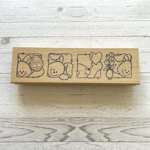 May include: A rectangular wooden stamp with four square designs. Each square features a cartoon bunny with an Easter egg, a cloud, a heart, and flowers. The bunnies are outlined in black.