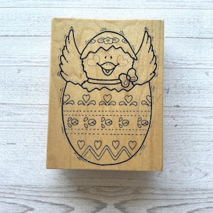 May include: A wooden stamp featuring a chick emerging from a decorated egg. The chick has wings and a bow. The egg is adorned with hearts, dotted lines, and zigzags. The stamp is on a light wood surface.