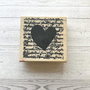 May include: A wooden stamp with a large black heart in the center. The background is filled with handwritten text in black ink. The stamp is square and sits on a light wood surface.