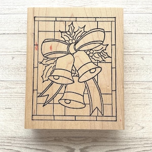 May include: A wooden stamp featuring a Christmas bell design. The stamp has a rectangular shape with a black outline of two bells, a bow, and holly leaves. The design is set against a grid pattern.