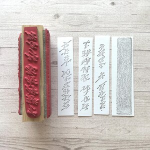 May include: A red rubber stamp with a wooden handle, and four rectangular paper strips with text. The stamp features a floral design and the text is in a script font. The paper strips have text and a textured pattern.