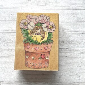May include: A wooden stamp featuring a whimsical illustration of a fairy in a flower pot. The fairy has long blonde hair and is surrounded by pink flowers and green leaves. The pot is decorated with hearts and a ladybug.