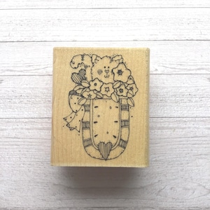 Puede incluir: Un sello de madera con un diseño caprichoso. El sello representa una criatura parecida a un oso con un ramo de flores y un lazo. El diseño está en tinta negra sobre un bloque de madera rectangular.