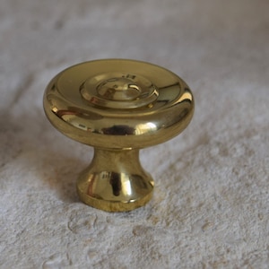 May include: A gold-toned cabinet knob with a round, slightly raised top and a smaller, centered circle. The knob has a short, cylindrical base.