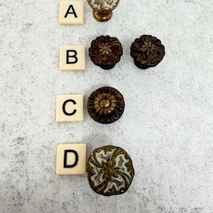 May include: A selection of decorative cabinet knobs in various styles. The knobs are in antique brass and bronze tones, with floral and ornate designs. Scrabble tiles with letters A, B, C, and D are placed next to each knob.