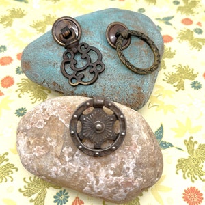 May include: Two antique brass cabinet knobs on a stone background. One knob is a round, ornate design with a ring handle. The other knob is a more intricate design with a decorative handle.