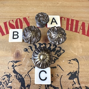 May include: Four ornate metal cabinet knobs with a floral design. The knobs are arranged in a circle with the letters A, B, and C on white tiles.