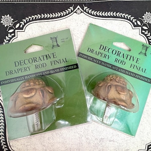 May include: Two decorative drapery rod finials in green packaging. The finials are bronze-colored and have a unique design. The packaging reads "DECORATIVE DRAPERY ROD FINIAL" and "INDIVIDUALLY HANDCAST AND HAND FINISHED".