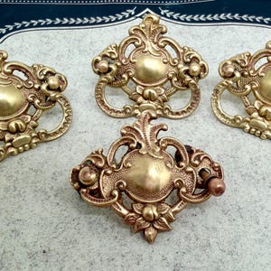 May include: Four antique brass drawer pulls with ornate, floral designs. The pulls have a warm, golden hue and feature intricate details, including a central medallion and curved handles. The hardware is likely for furniture restoration or decorative projects.