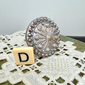 Może przedstawiać: Ozdobny, srebrny uchwyt do szafki o okrągłym wzorze z wypukłymi kropkami i centralnym wzorem w kształcie gwiazdy. Obok uchwytu znajduje się kremowa płytka z literą "D". Uchwyt leży na białej koronkowej serwetce.