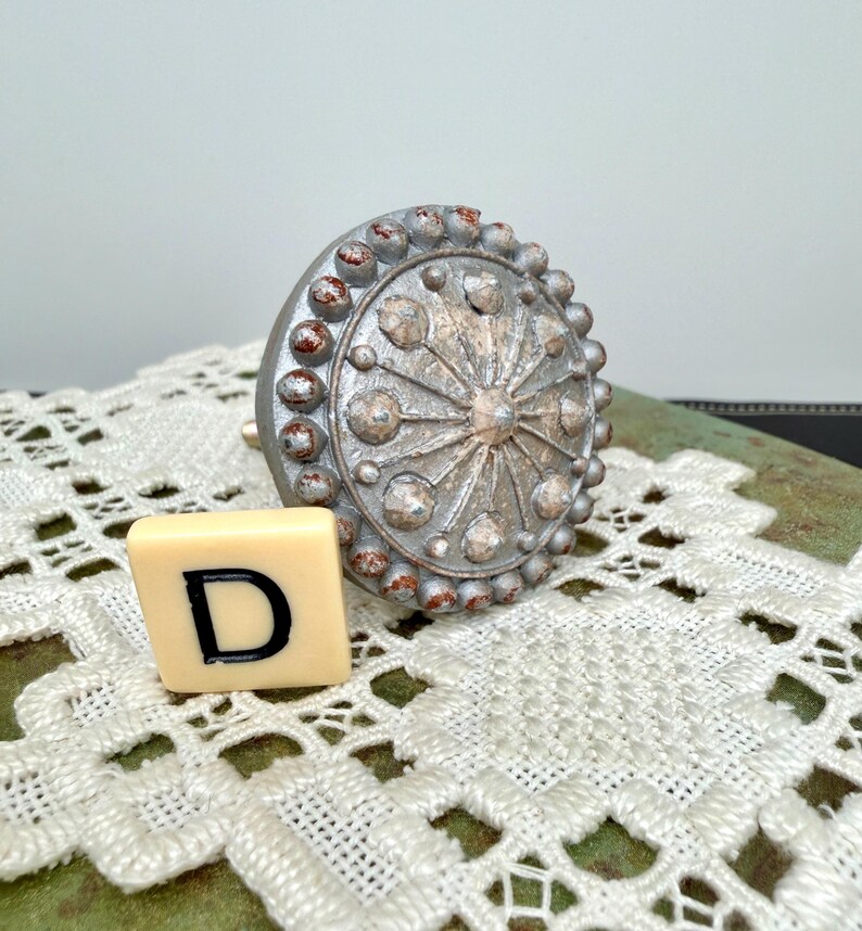 Może przedstawiać: Ozdobny, srebrny uchwyt do szafki o okrągłym wzorze z wypukłymi kropkami i centralnym wzorem w kształcie gwiazdy. Obok uchwytu znajduje się kremowa płytka z literą "D". Uchwyt leży na białej koronkowej serwetce.