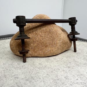 May include: A dark brown metal furniture handle with a horizontal bar and vertical posts with threaded rods. The handle is mounted on a light brown rock, set against a neutral background.