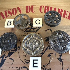 May include: Five antique-style metal drawer pulls with intricate designs. The pulls are arranged in a circle on a wooden surface with letters A, B, C, D, and E surrounding them.
