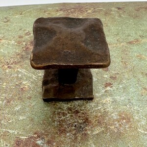 Puede incluir: Un tirador de armario cuadrado de color bronce con una superficie texturizada. El tirador tiene una base rectangular y una parte superior cuadrada con bordes redondeados. El herraje es probablemente de metal y est&aacute; dise&ntilde;ado para su uso en un armario o caj&oacute;n.