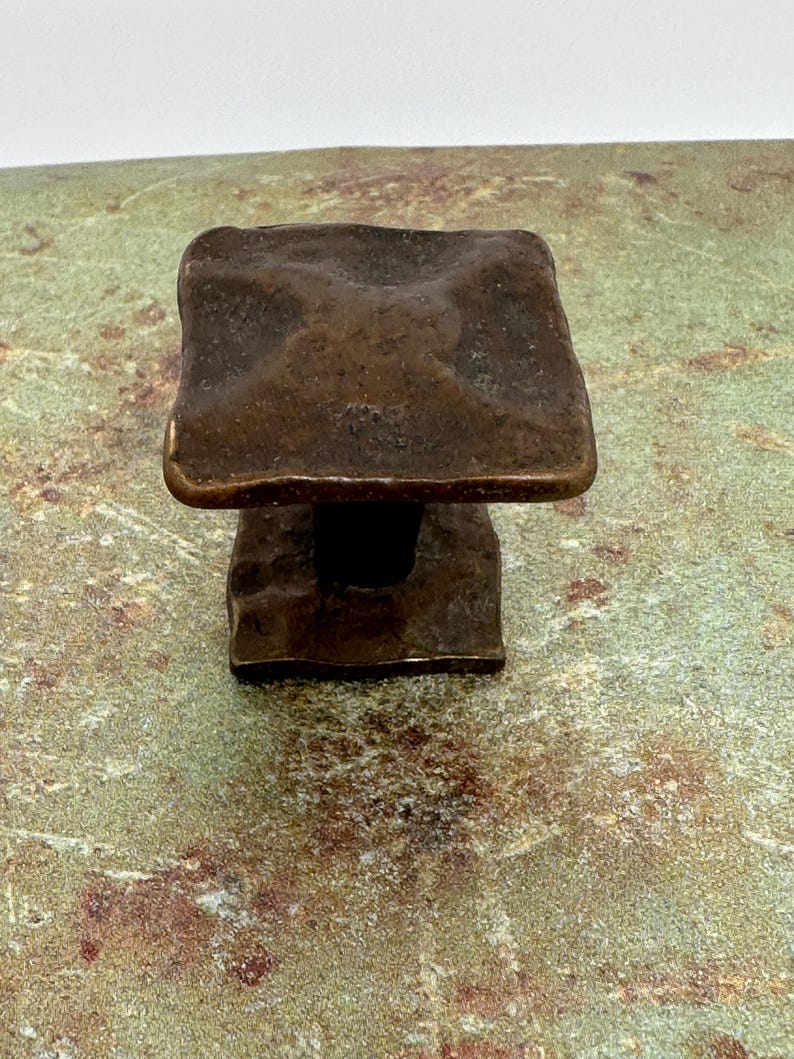 Puede incluir: Un tirador de armario cuadrado de color bronce con una superficie texturizada. El tirador tiene una base rectangular y una parte superior cuadrada con bordes redondeados. El herraje es probablemente de metal y est&aacute; dise&ntilde;ado para su uso en un armario o caj&oacute;n.