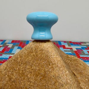 May include: A light blue ceramic knob with a rounded top and a flared base sits atop a textured, brown, rock-like form. The background features a colorful, geometric pattern.