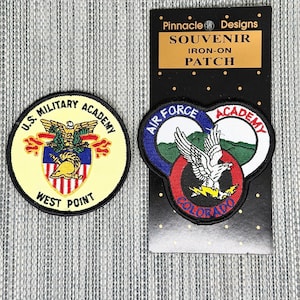 May include: Two embroidered patches. One is a circular U.S. Military Academy West Point patch with a gold helmet and eagle. The other is an Air Force Academy Colorado patch on a Pinnacle Designs card labeled SOUVENIR IRON-ON PATCH.