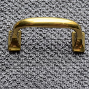 May include: A gold-toned metal handle with a rounded top and square bases. The handle is on a gray textured surface.