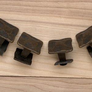 Puede incluir: Cuatro tiradores de armario de color bronce con un dise&ntilde;o rectangular texturizado. Cada tirador tiene un v&aacute;stago corto y una base cuadrada. Los tiradores est&aacute;n dispuestos sobre una superficie de madera clara.