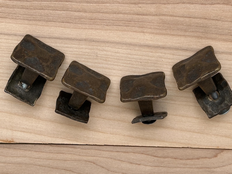 Puede incluir: Cuatro tiradores de armario de color bronce con un dise&ntilde;o rectangular texturizado. Cada tirador tiene un v&aacute;stago corto y una base cuadrada. Los tiradores est&aacute;n dispuestos sobre una superficie de madera clara.