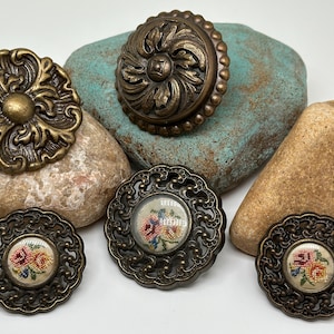 May include: An assortment of antique bronze-toned decorative hardware. The collection includes ornate drawer pulls and decorative buttons with floral needlepoint centers. The hardware is displayed on natural stones, showcasing their vintage aesthetic.