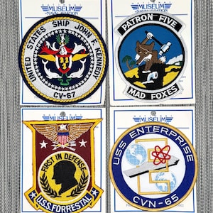 May include: Four embroidered military patches on white backing cards. Top left: USS John F. Kennedy CV-67 circular patch. Top right: Patron Five Mad Foxes patch with a fox. Bottom left: USS Forrestal First in Defense shield. Bottom right: USS Enterprise CVN-65 patch.