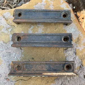 May include: Three antique metal door hinges with a rusty finish. The hinges are arranged in a row on a rock surface.