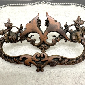 May include: Ornate bronze-colored drawer pull with a curved handle and decorative floral and leaf accents. The pull features a symmetrical design with detailed scrollwork and a vintage aesthetic. The hardware is likely for a cabinet or dresser.