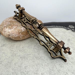 May include: An antique bronze-toned door pull with an ornate design. The pull features a long, rectangular backplate with decorative cutouts and a central handle with a geometric shape. The hardware is likely made of metal and has a vintage aesthetic.