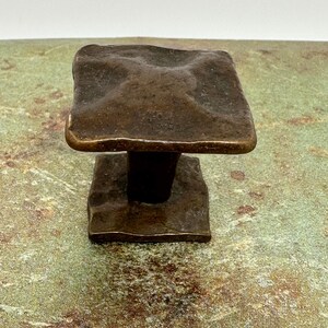 Puede incluir: Un tirador de armario cuadrado de color bronce con una superficie texturizada. Tiene una base cuadrada m&aacute;s peque&ntilde;a y un v&aacute;stago cil&iacute;ndrico corto que conecta los dos cuadrados. El acabado tiene un aspecto r&uacute;stico y envejecido.