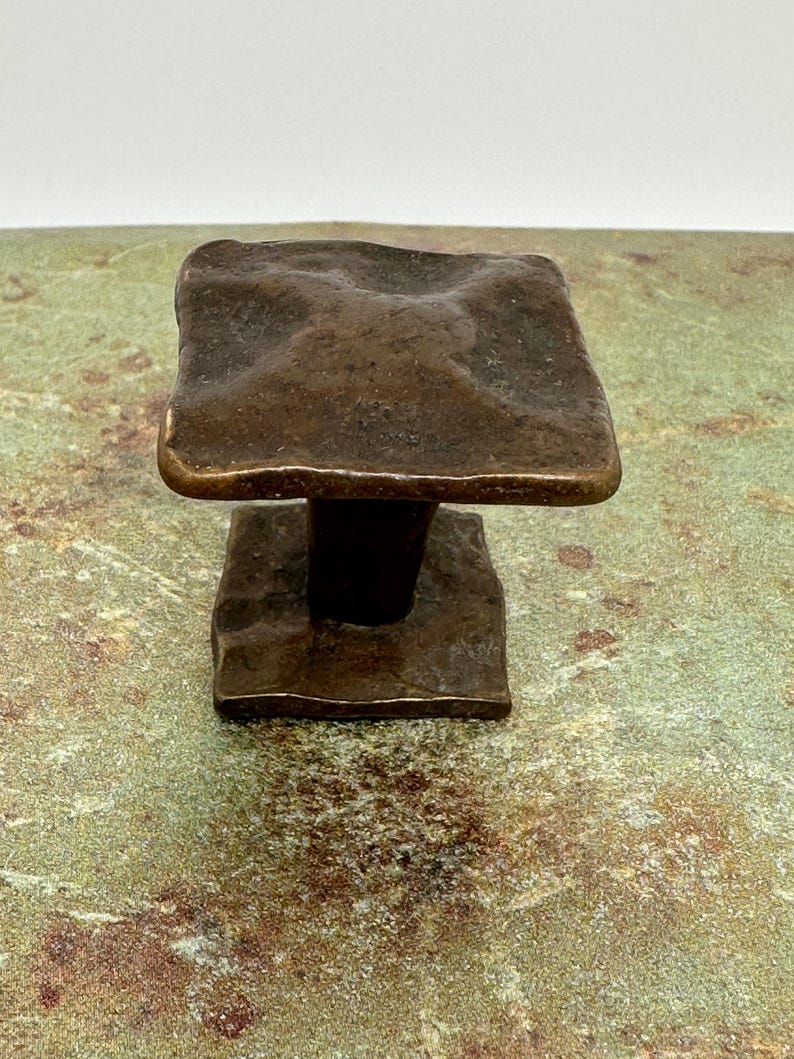 Puede incluir: Un tirador de armario cuadrado de color bronce con una superficie texturizada. Tiene una base cuadrada m&aacute;s peque&ntilde;a y un v&aacute;stago cil&iacute;ndrico corto que conecta los dos cuadrados. El acabado tiene un aspecto r&uacute;stico y envejecido.